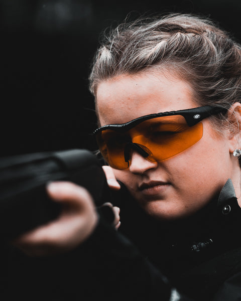 Woman aiming a shotgun in black Methow frames with orange lenses; lifestyle colorway
