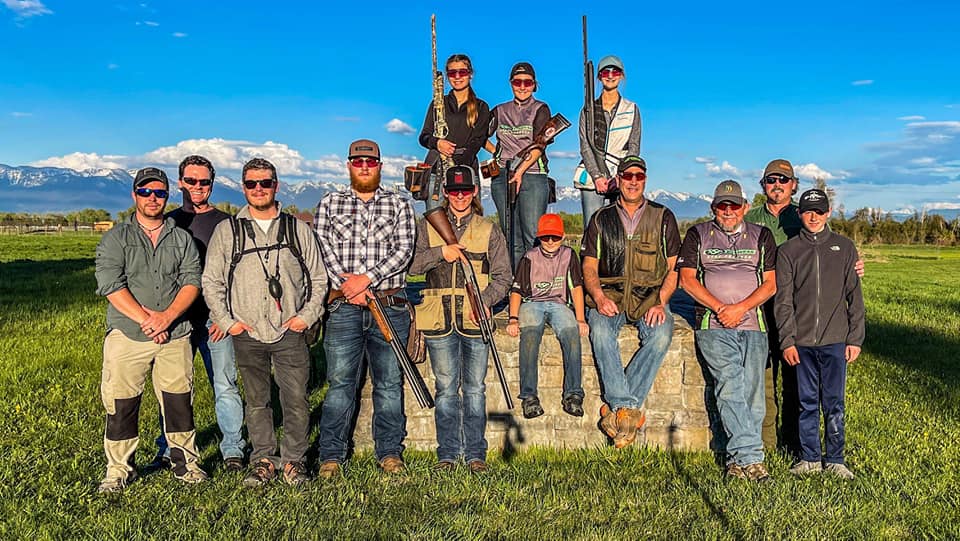 Youth and adult shooters wearing eyewear together outdoors, with mountains behind them.