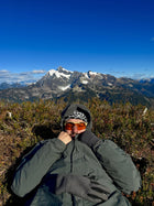 Roark facing the camera in orange-lens Methow eyewear, holding his hood with mountain peaks behind him
