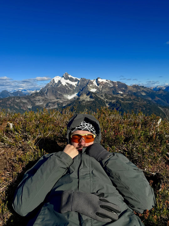 Roark facing the camera in orange-lens Methow eyewear, holding his hood with mountain peaks behind him